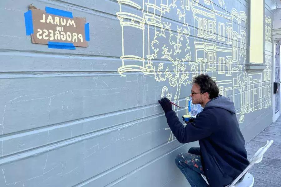 一位艺术家在教会区一栋建筑的一侧画了一幅壁画, 建筑物上贴着“正在画壁画”的牌子. 贝博体彩app，加利福尼亚州.
